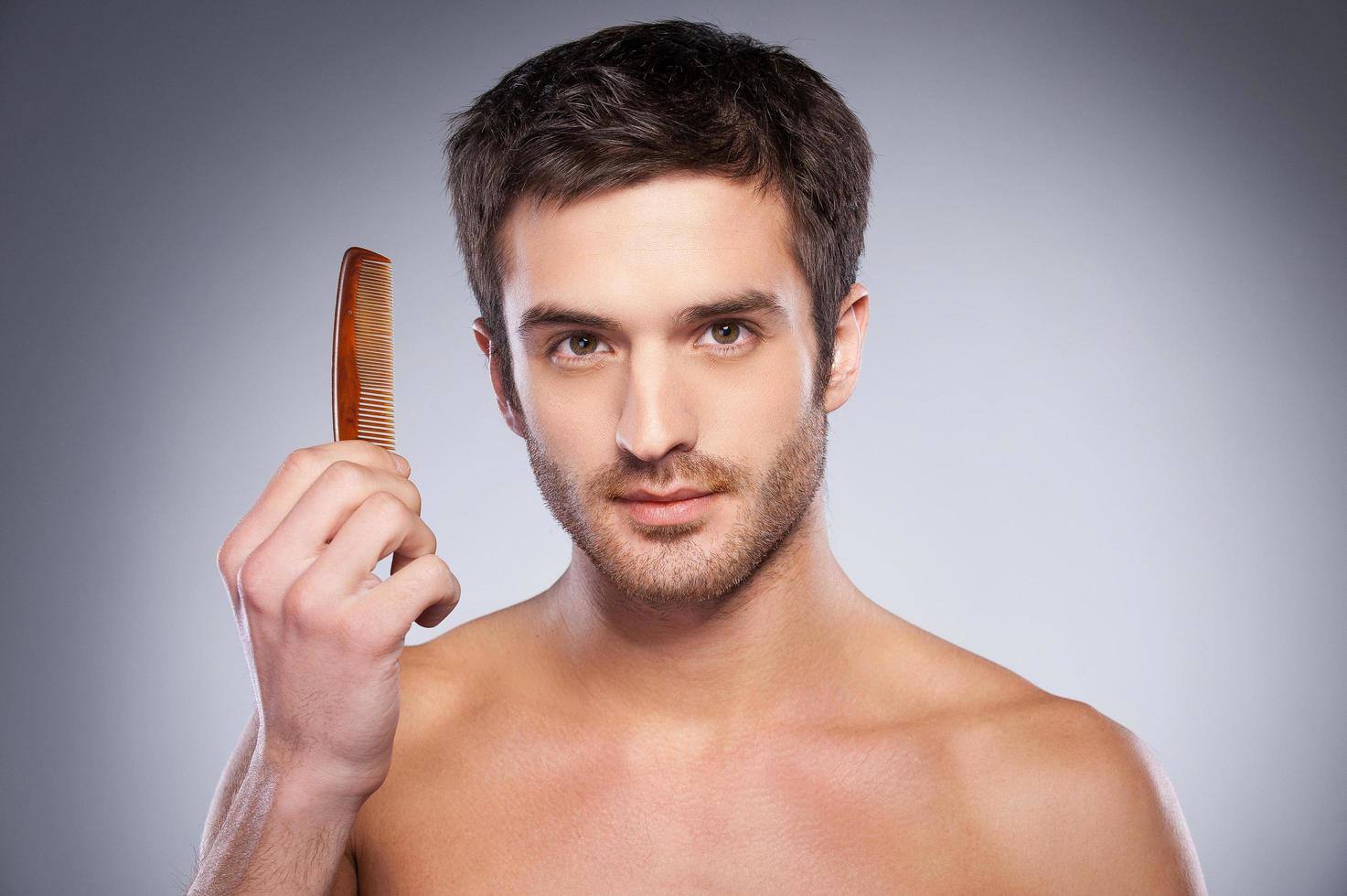 Making his unique style. Handsome young shirtless man holding a comb and looking at camera while standing against grey background photo