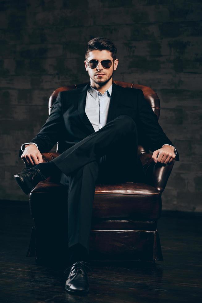 elegancia y masculinidad. joven apuesto hombre de traje y gafas de sol con la mano en la barbilla y mirando a la cámara mientras está sentado en una silla de cuero contra un fondo gris oscuro foto