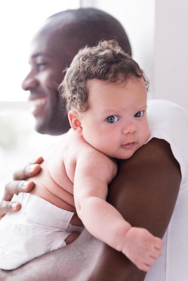 Papá orgulloso y feliz. feliz joven africano sosteniendo a su pequeño bebé y sonriendo mientras está de pie cerca de la ventana foto