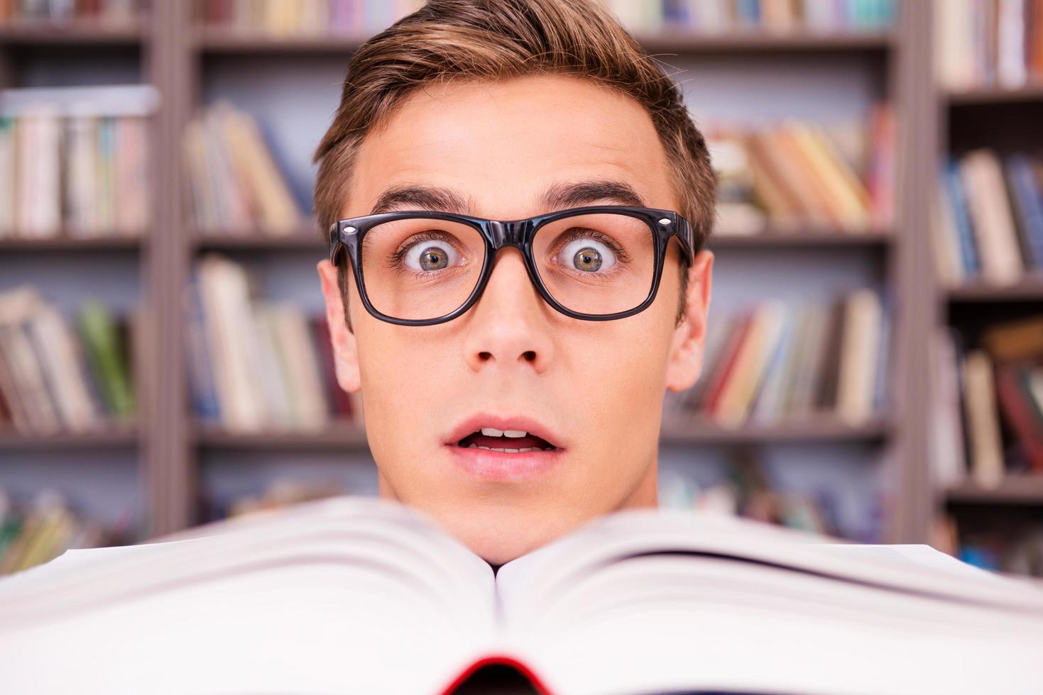 Surprised bookworm. Surprised young man looking out of the book and keeping mouth open with bookshelf in the background photo