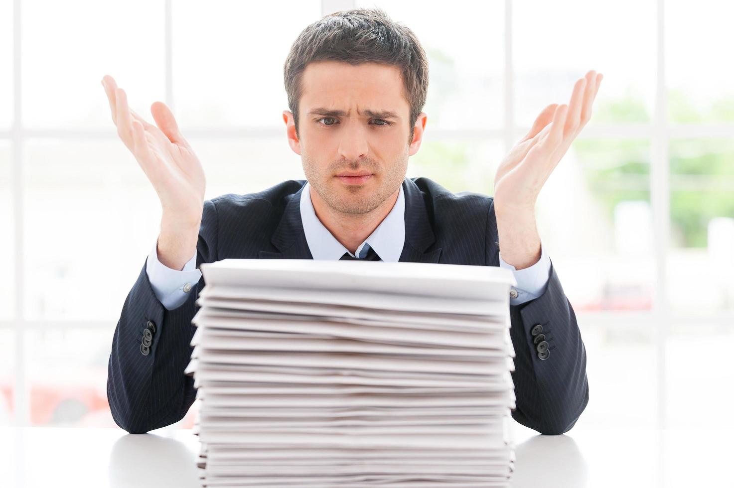 Hopeless businessman. Depressed young man in shirt and tie looking at the stack of paperwork and gesturing while sitting at his working place photo