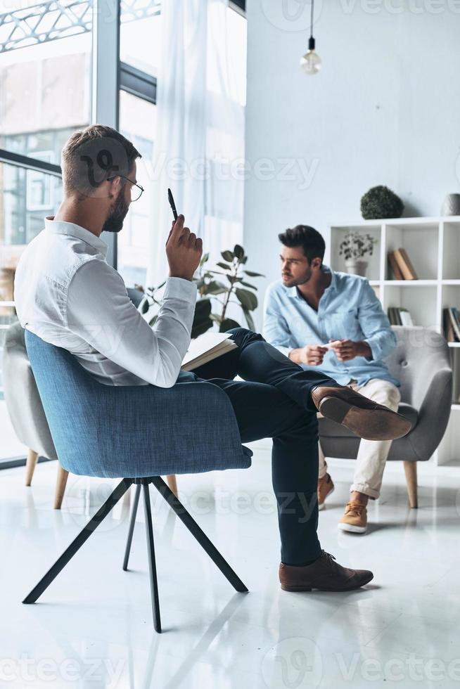Always hard to speak out. Young frustrated man solving his mental problems while having therapy session with psychologist photo