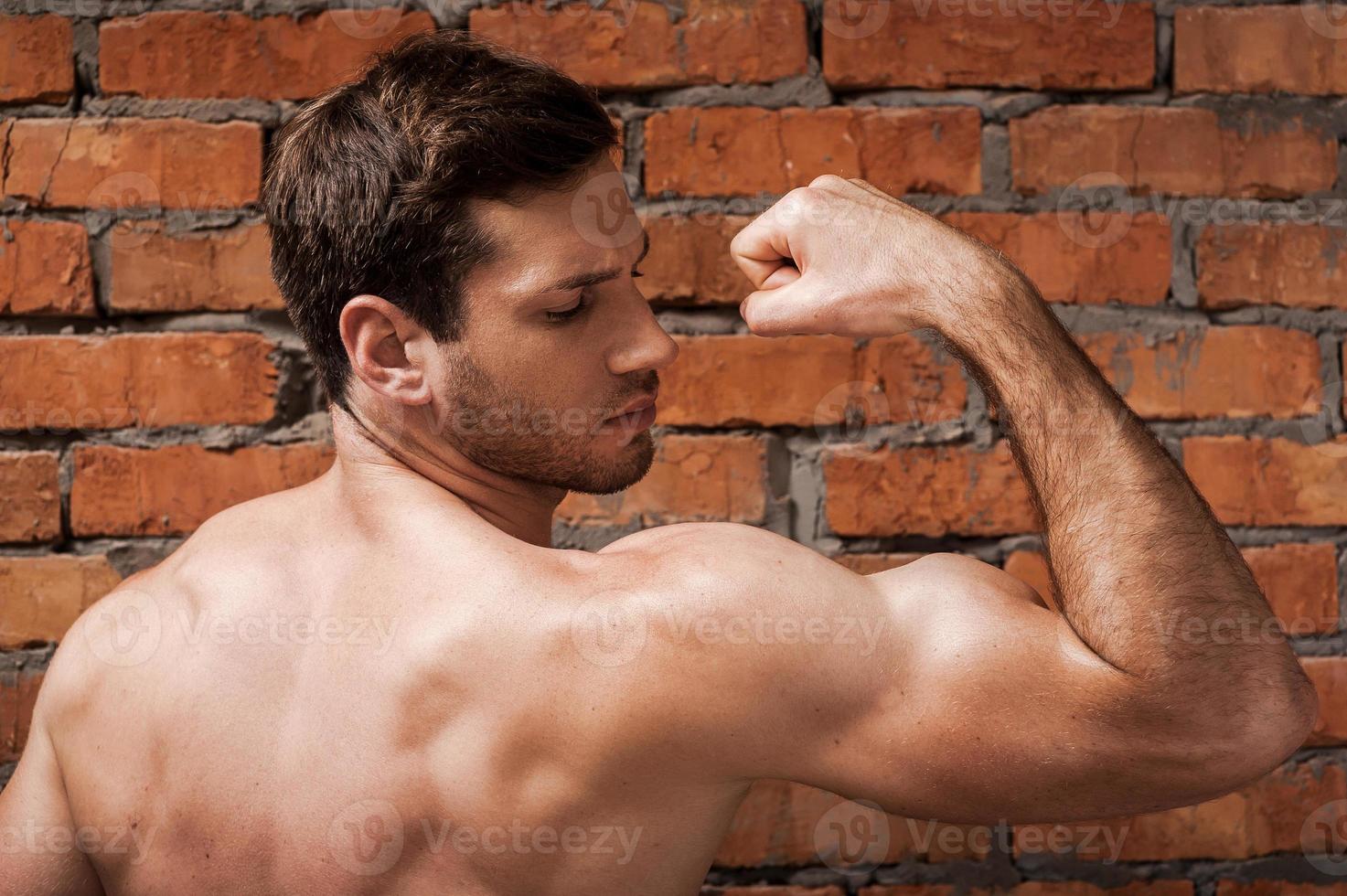 Showing his perfect bicep. Rear view of confident young muscular man posing while standing against brick wall photo