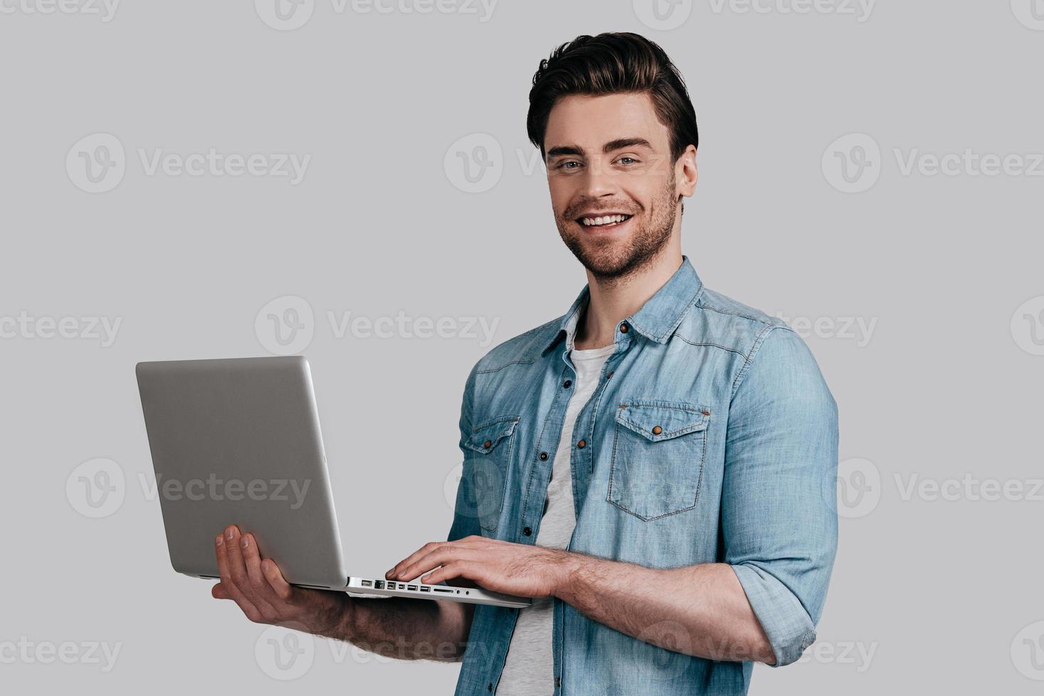 May I help you Handsome young man in blue jeans shirt holding laptop and looking at camera while standing against grey background photo