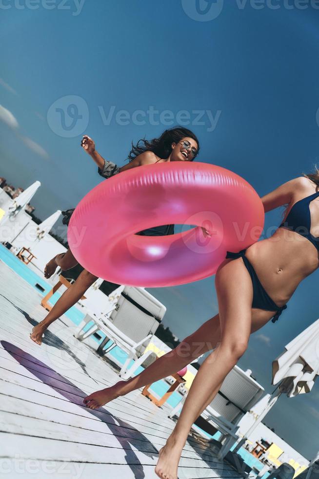sentirse libre y feliz. dos hermosas mujeres jóvenes en bikini corriendo juntas al aire libre mientras una de ellas lleva un anillo inflable foto
