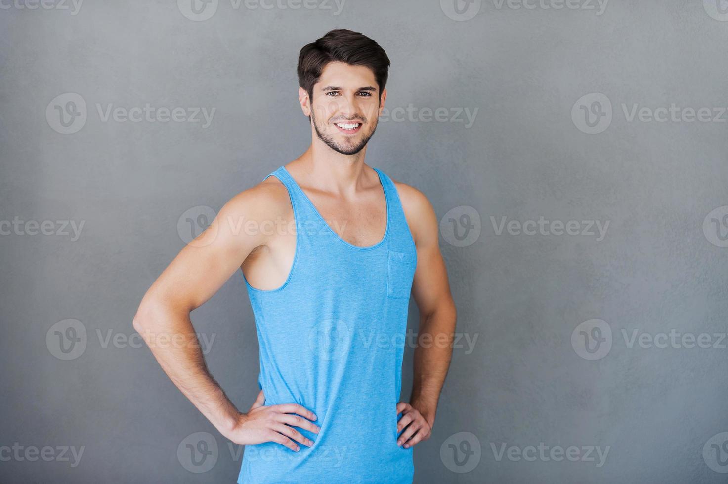 Fit and confident. Happy young muscular man holding hands on hip while standing against grey background photo