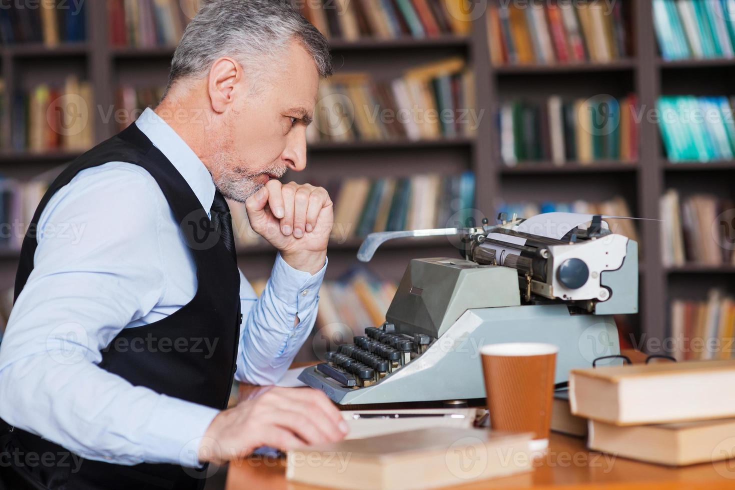 Waiting for inspiration. Side view of confident grey hair senior man in formalwear sitting at the typewriter and holding hand on chin with bookshelf in the background photo