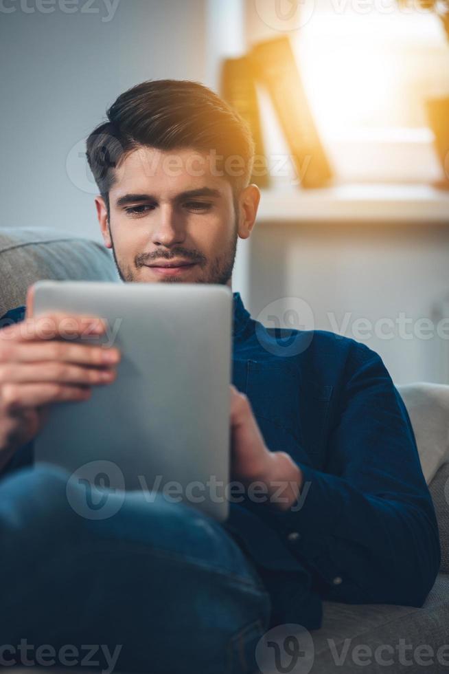 Enjoying his new digital tablet. Close-up of handsome young man using his digital tablet with smile while lying down on the couch at home photo