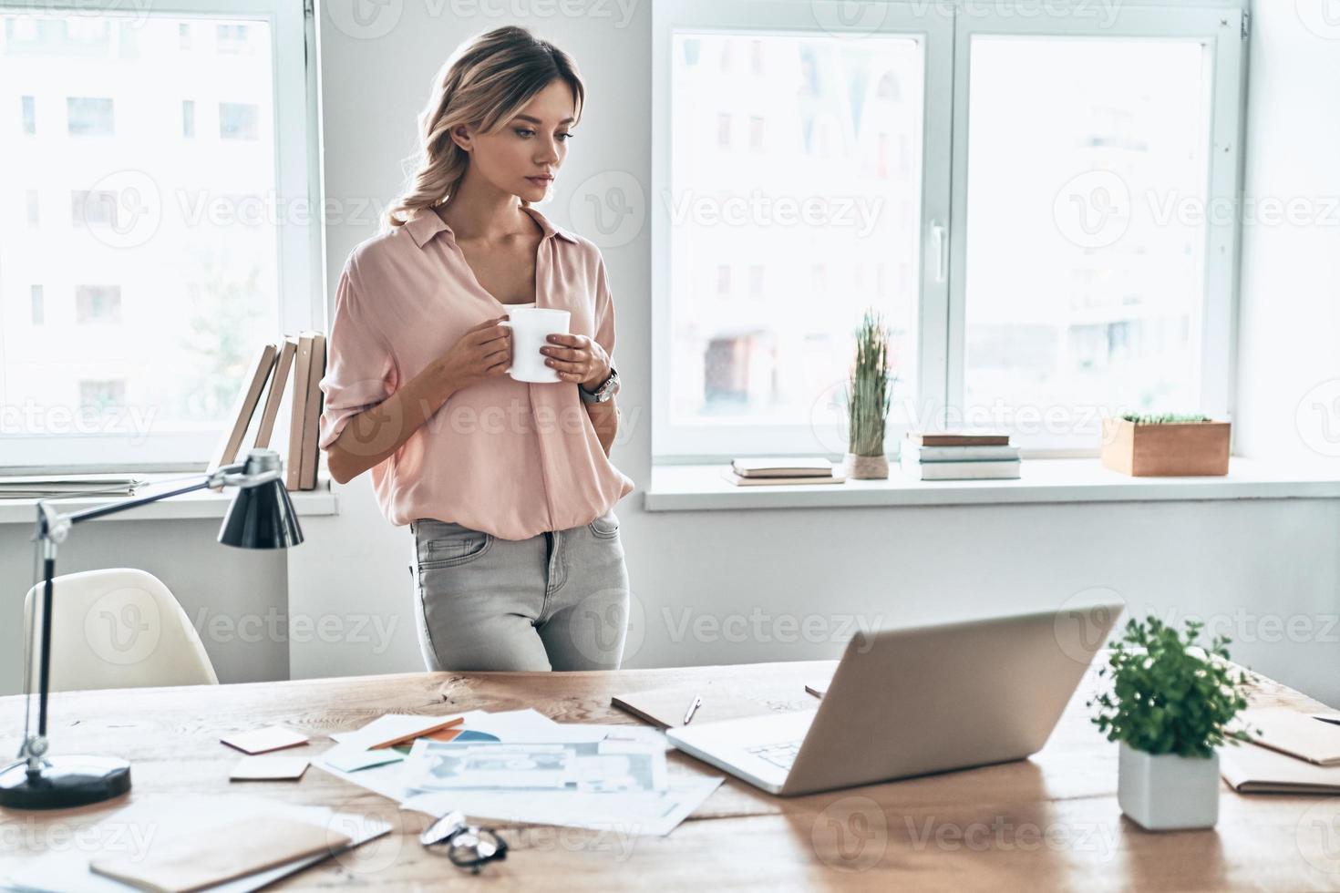 nueva solución cada día. mujer joven con ropa informal elegante sosteniendo una taza de café y mirando una laptop mientras está pie en una oficina moderna 13525793 Foto de stock