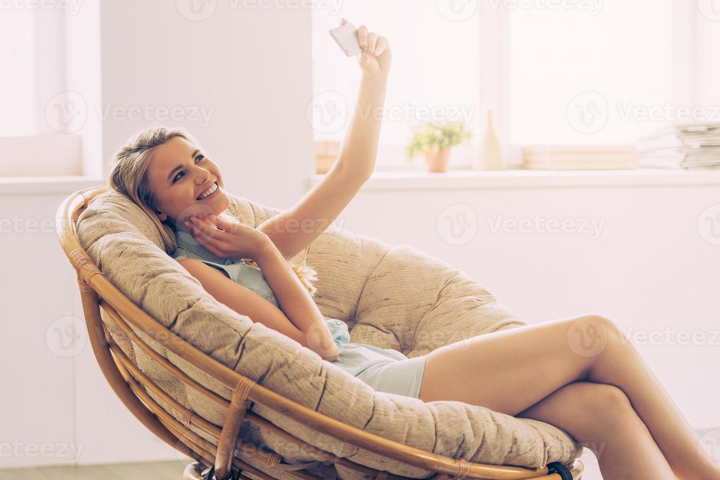 tiempo de selfie hermosa joven haciendo selfie con su teléfono inteligente y sonriendo mientras se sienta en una silla grande y cómoda en casa foto