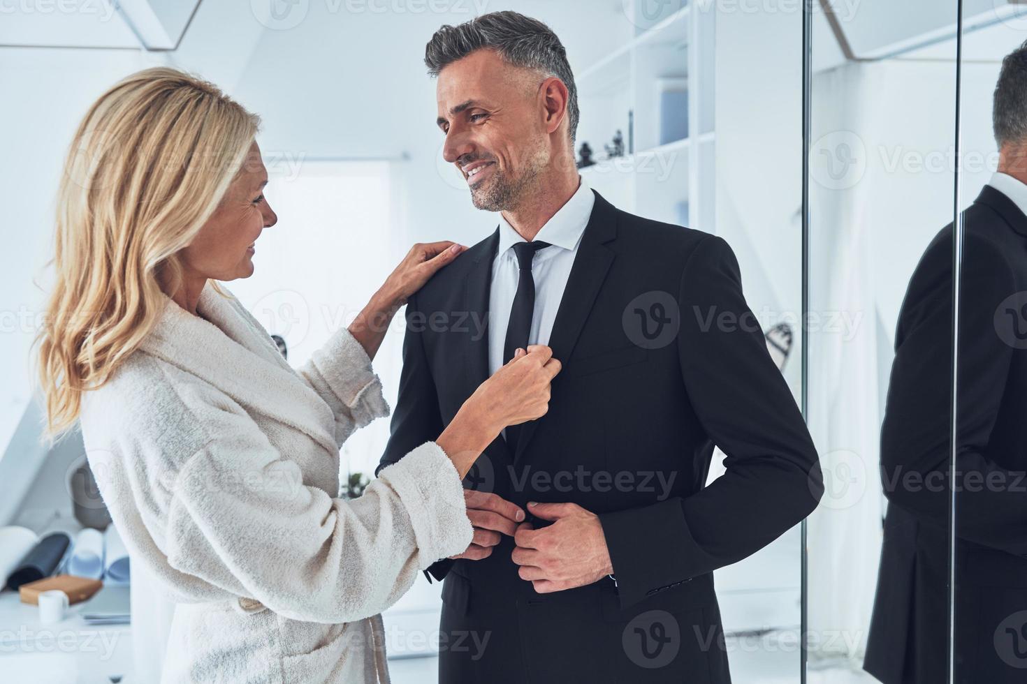 Beautiful mature woman adjusting necktie of her husband while both standing in front of the mirror at home photo