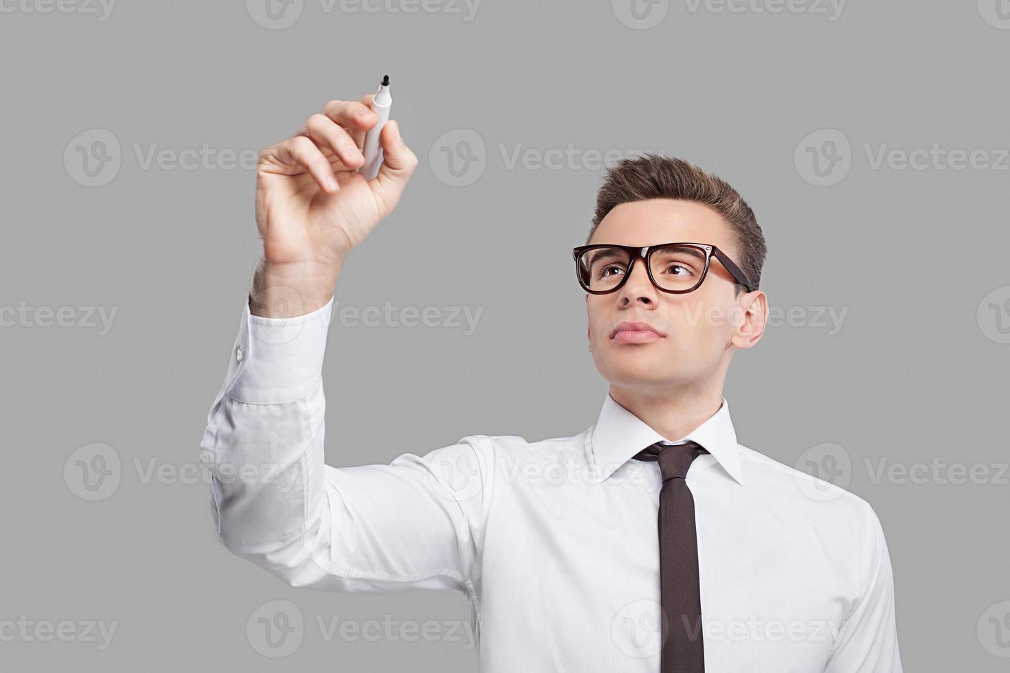 Transparent wipe board. Thoughtful young man in shirt and tie writing on the transparent wipe board while standing against grey background photo