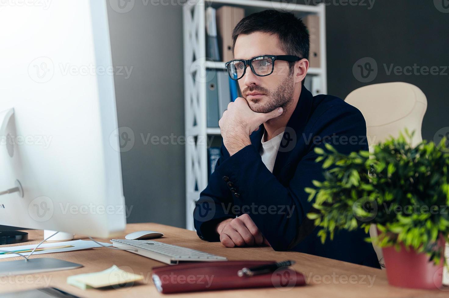 Thinking about solution. Thoughtful young man in smart casual wear holding hand on chin and looking at computer monitor while sitting at his working place in office photo
