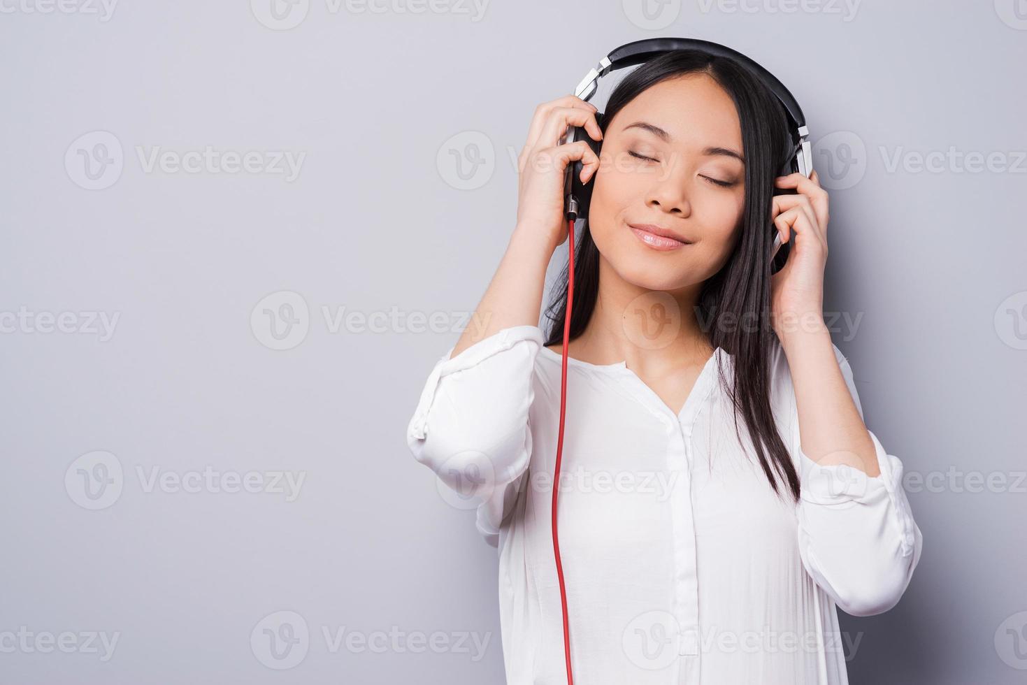 Music waves. Beautiful young Asian in headphones keeping eyes closed and smiling while standing against grey background photo