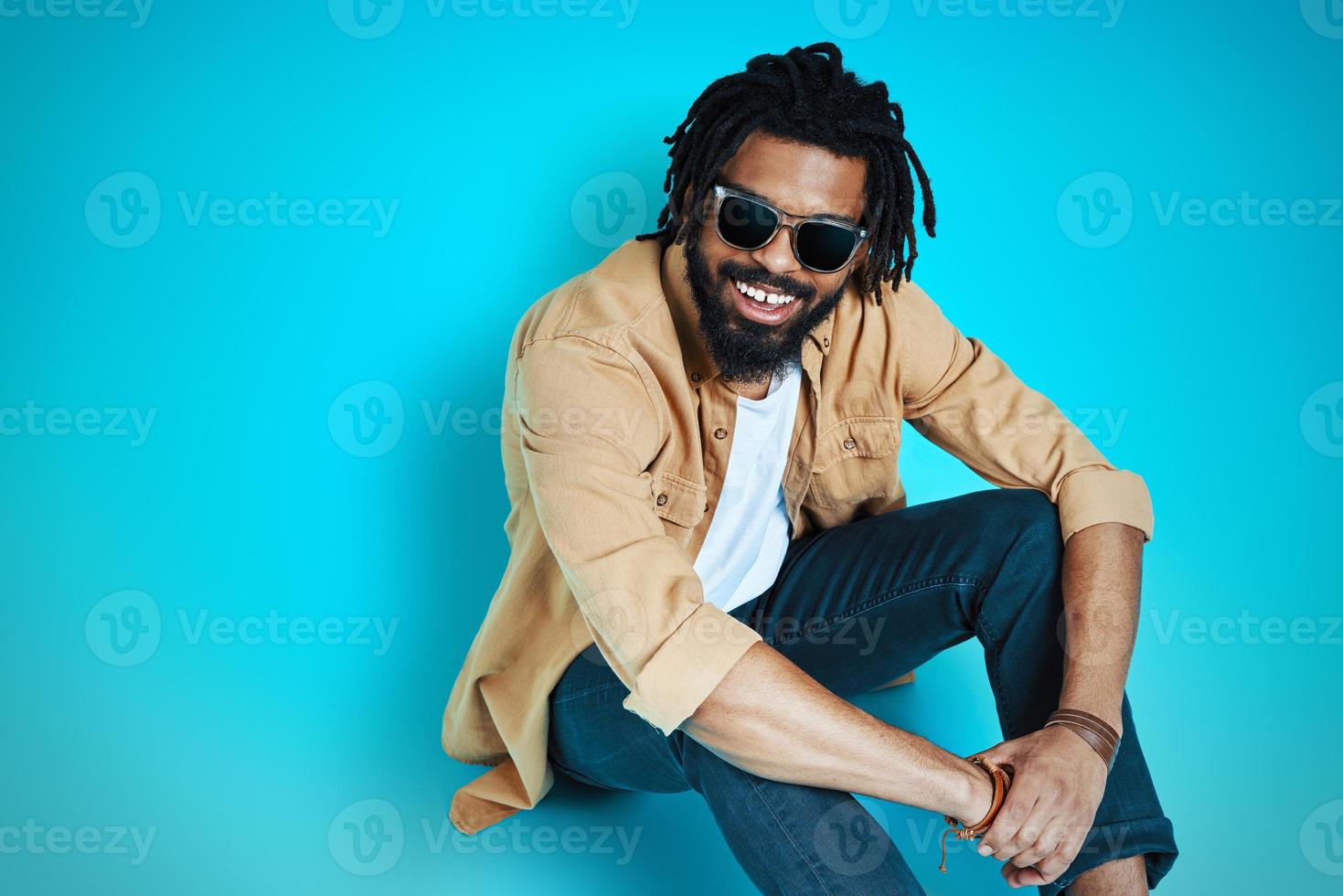 Top view of charming young African man in casual wear smiling and looking at camera while sitting against blue background photo