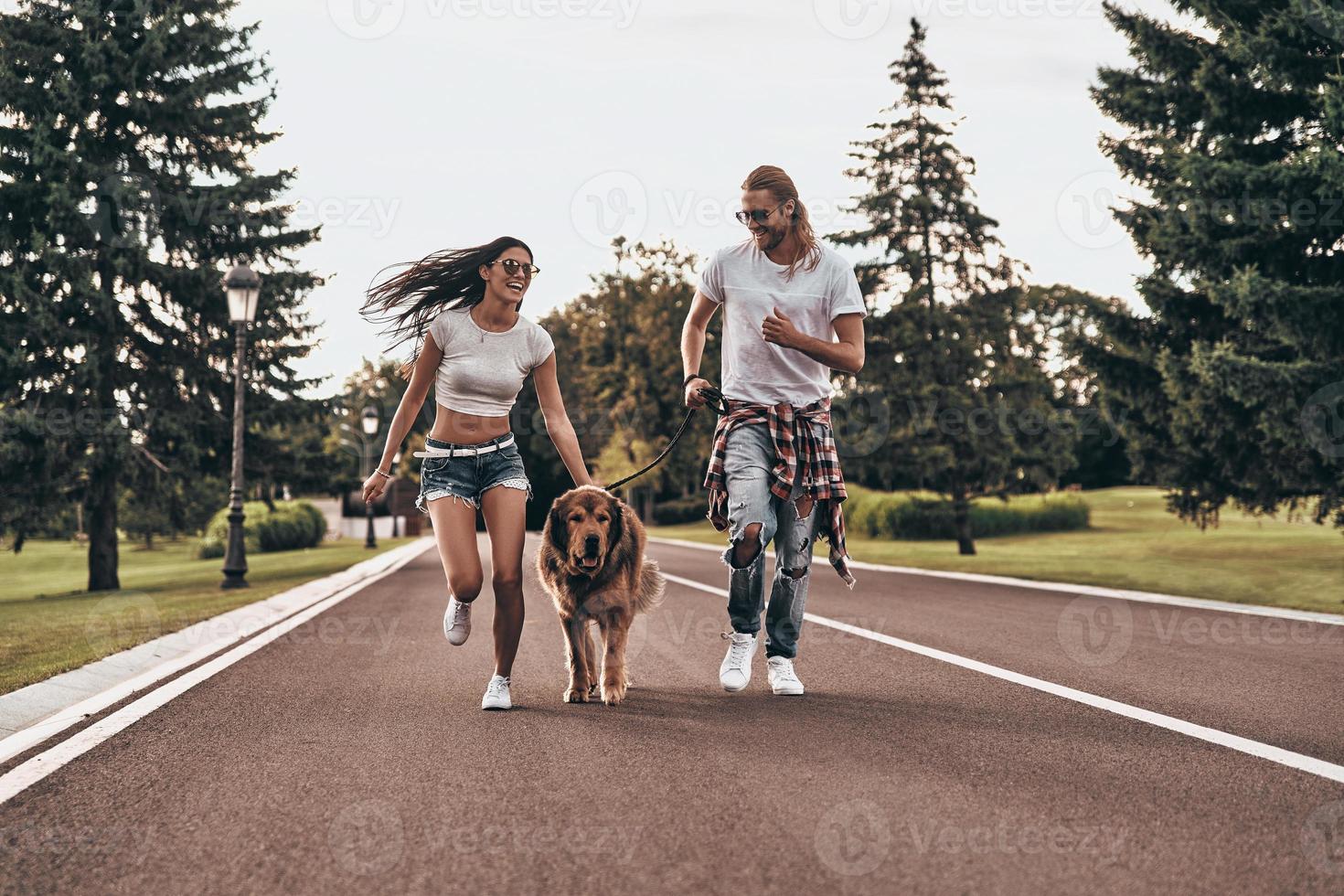 Nice day in park. Full length of beautiful young couple running with their dog while spending time outdoors photo