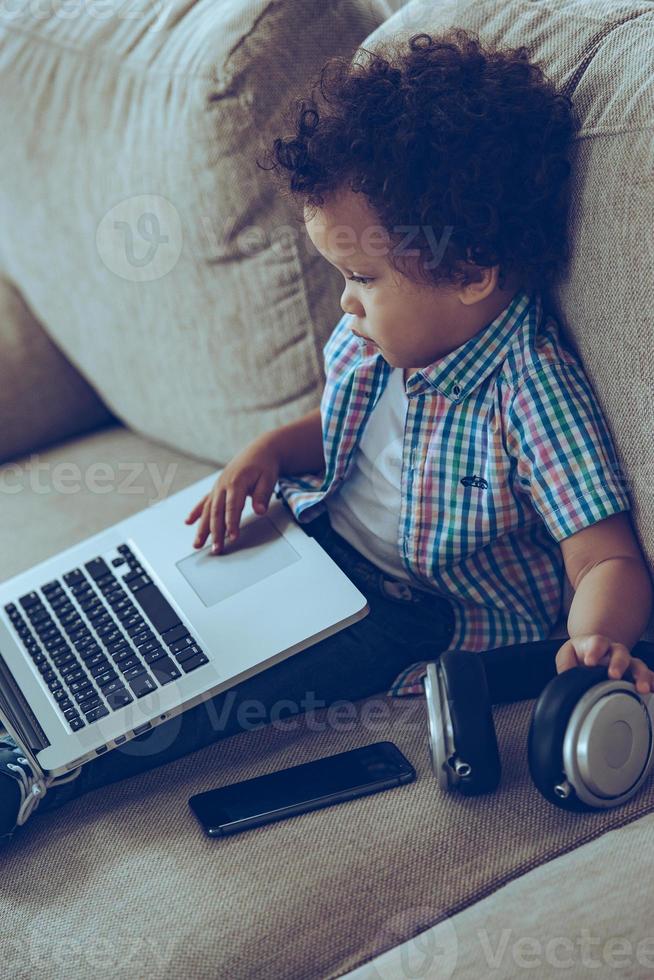 Little expert. Side view of little African baby boy using laptop while sitting on the couch at home photo