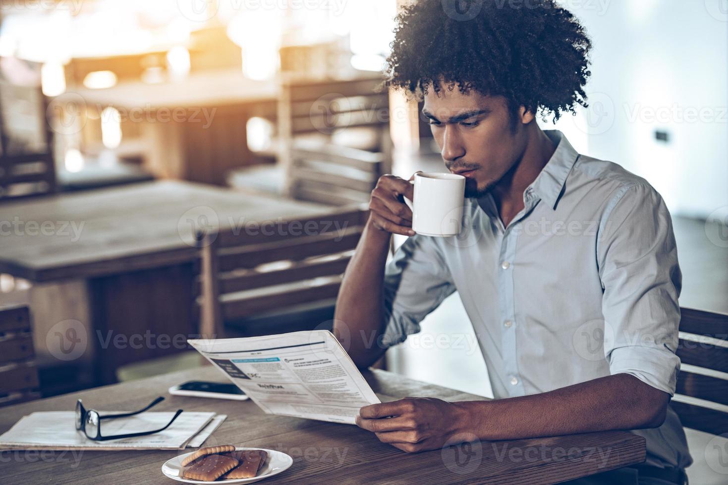 Comenzando nuevo gran día. joven africano leyendo el periódico y bebiendo café mientras está sentado en el café foto