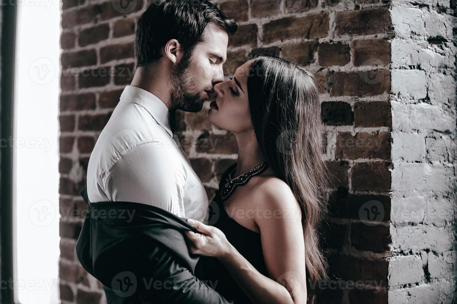 Stealing a kiss. Close-up side view of beautiful young woman in cocktail dress kissing her boyfriend and taking off his jacket while standing near brick wall in loft interior photo