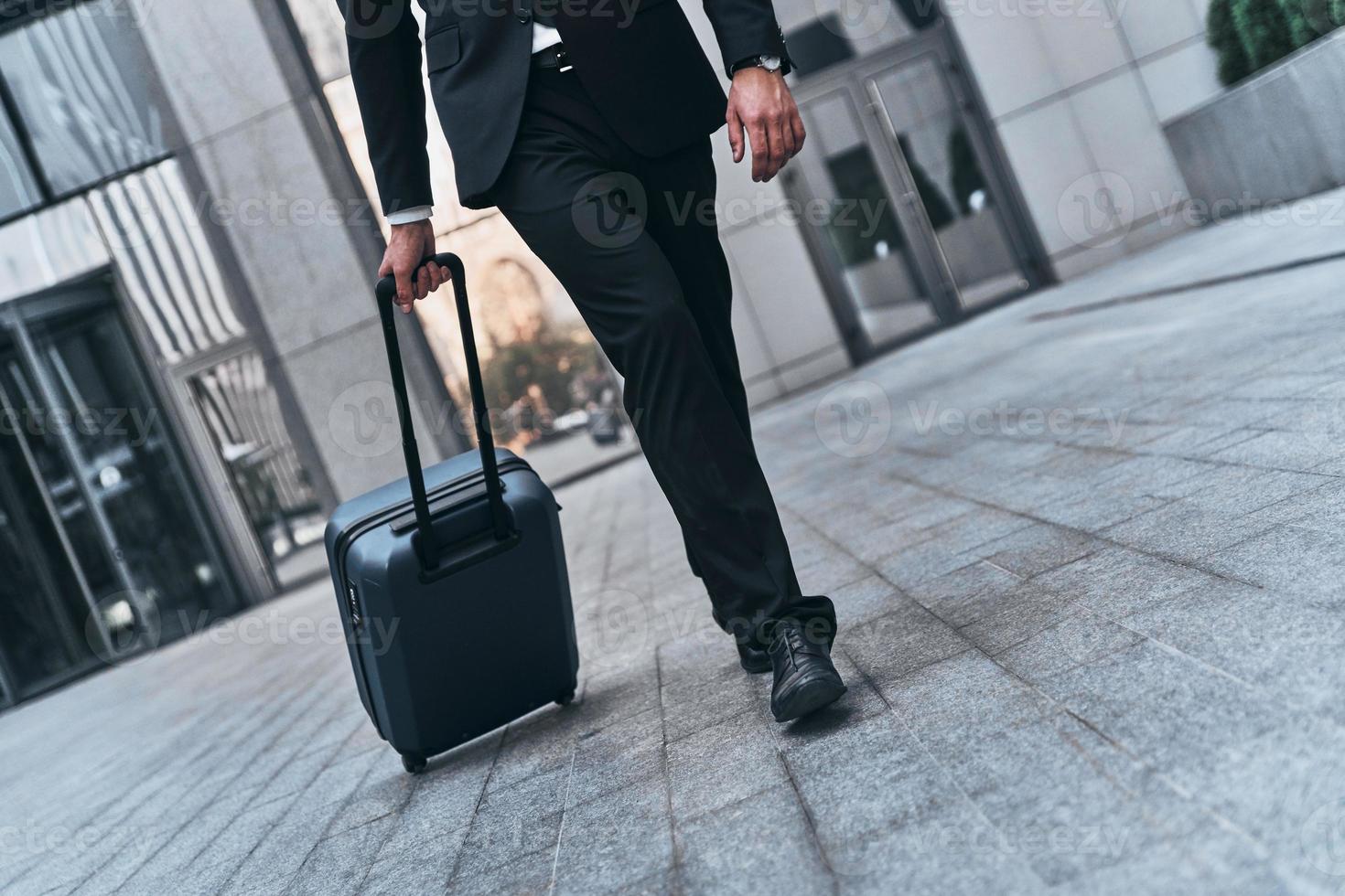 Business travel. Close up of man in full suit pulling luggage while walking outdoors photo