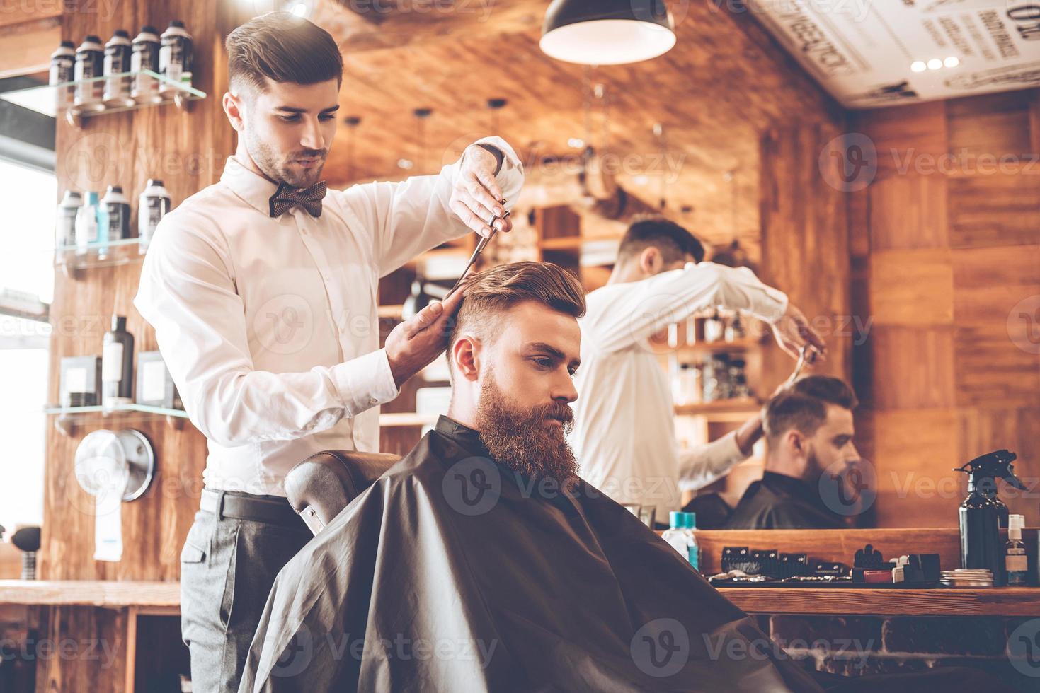 Everything should be perfect. Side view of young bearded man getting haircut by hairdresser while sitting in chair at barbershop photo