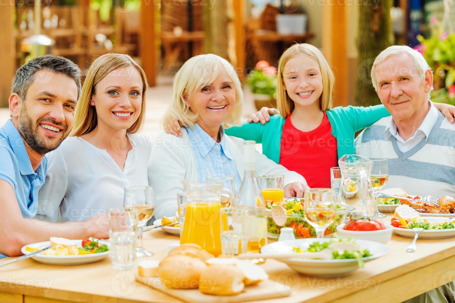 reunión familiar. familia feliz de cinco personas que se unen y sonríen mientras se sientan en la mesa del comedor al aire libre foto