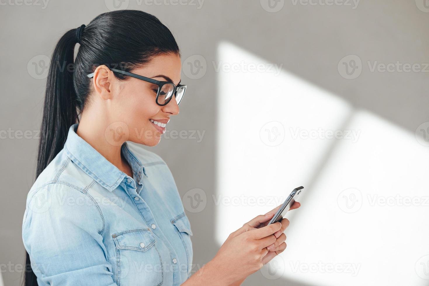 usando su teléfono inteligente con placer. vista lateral de primer plano de una hermosa joven alegre con anteojos usando su teléfono inteligente con una sonrisa mientras se enfrenta a un fondo gris foto