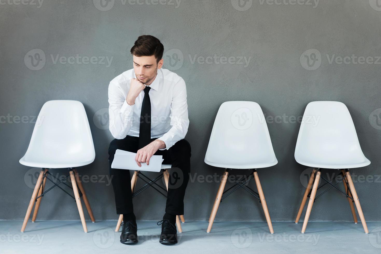 Long time of waiting. Pensive young businessman holding paper and holding hand on chin while sitting on chair against grey background photo