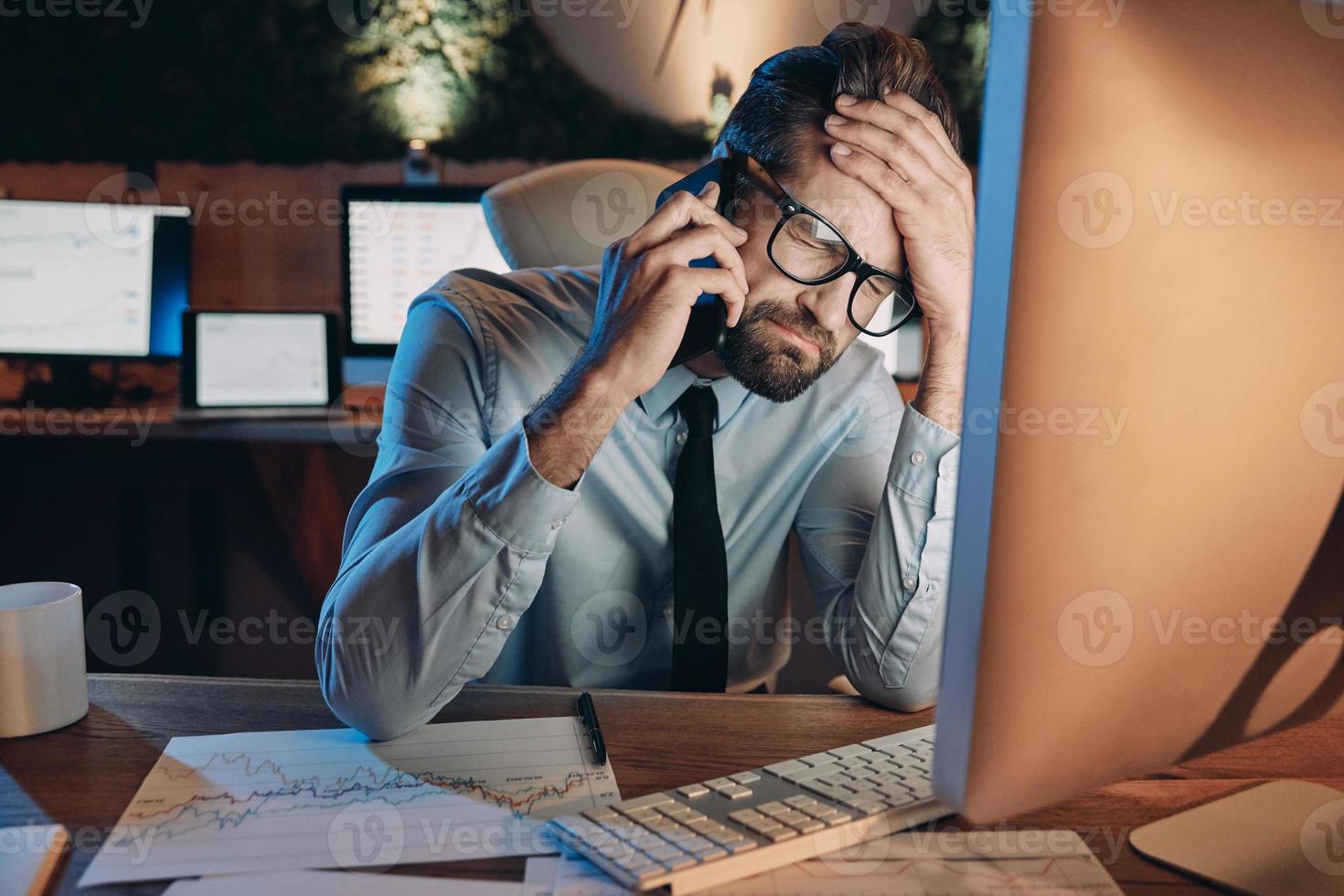 joven frustrado hablando por teléfono móvil y manteniendo los ojos cerrados mientras se queda tarde en la oficina foto