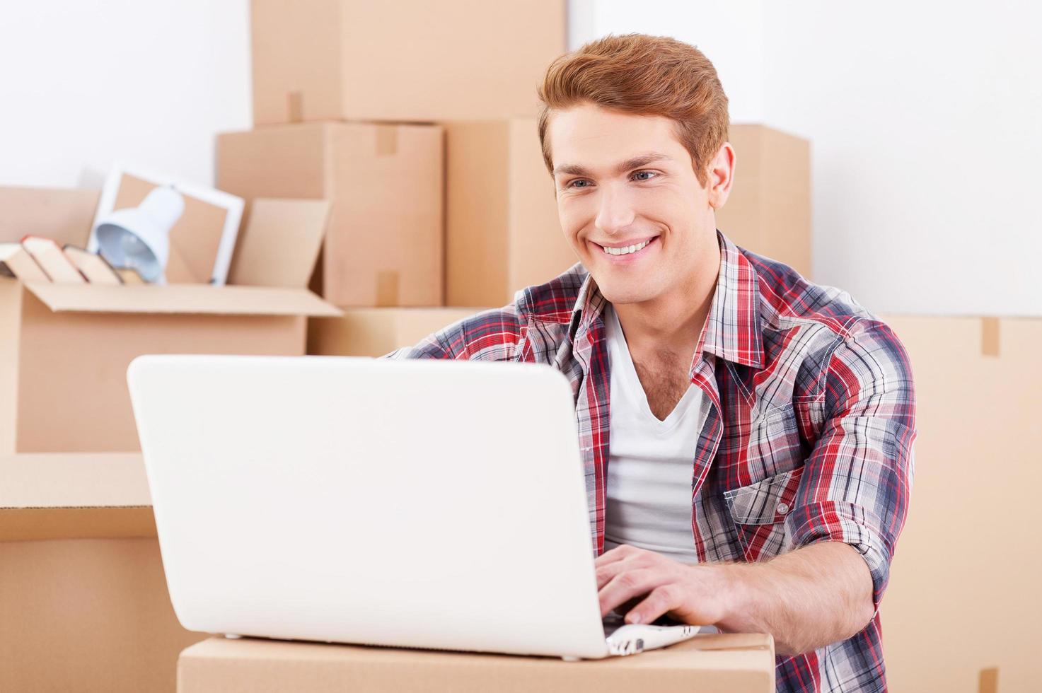 In search of a good moving company. Handsome young man sitting on the floor and working on laptop while cardboard boxes laying in the background photo