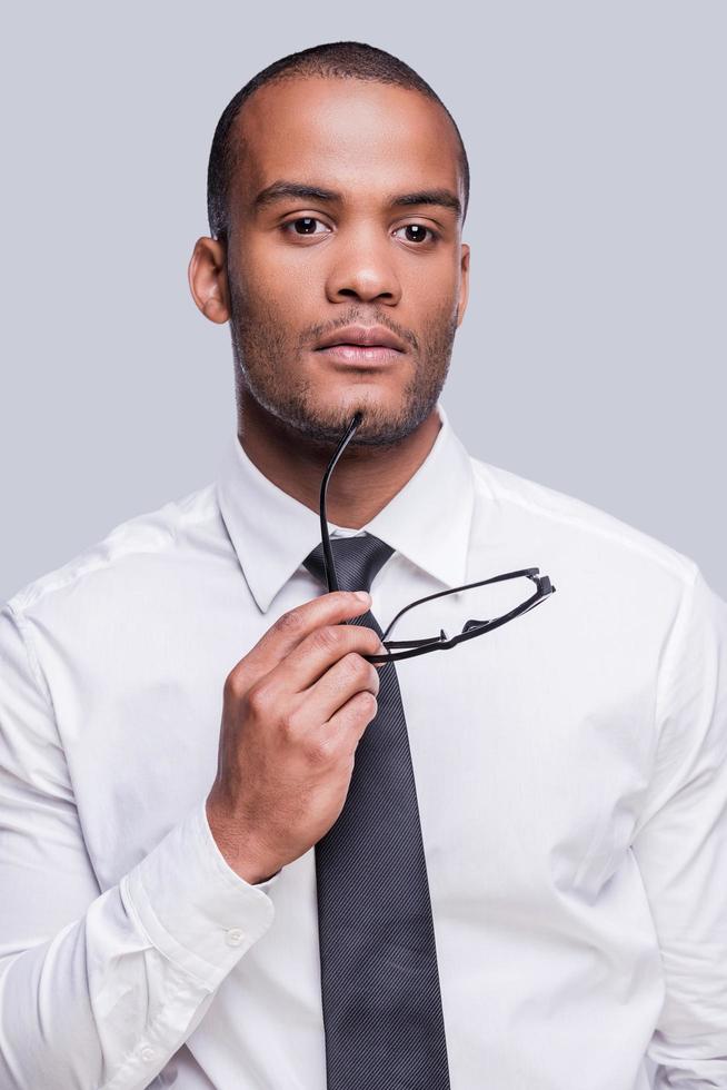 Looking  for inspiration. Thoughtful young African man in shirt and tie holding eyeglasses and looking away while standing against grey background photo