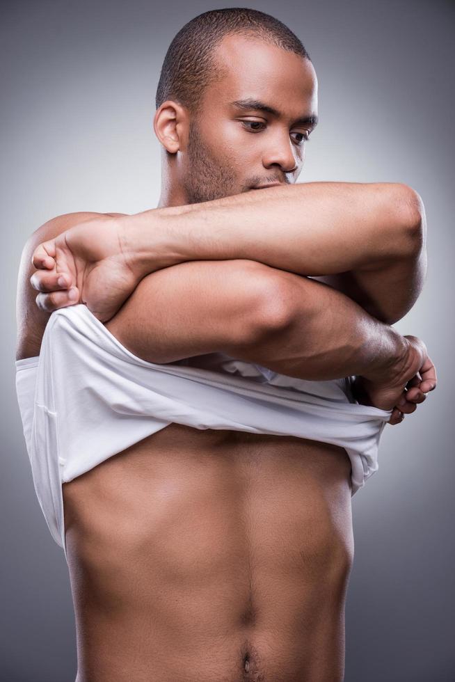 Taking his clothes away. Handsome young African man taking off his tank top while standing against grey background photo