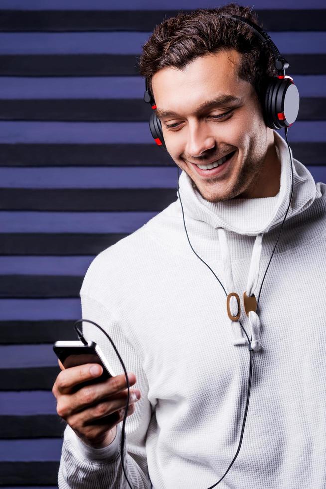 elegir música para escuchar. un joven apuesto con auriculares sosteniendo un reproductor de mp3 y mirándolo con una sonrisa mientras se enfrenta a un fondo rayado foto