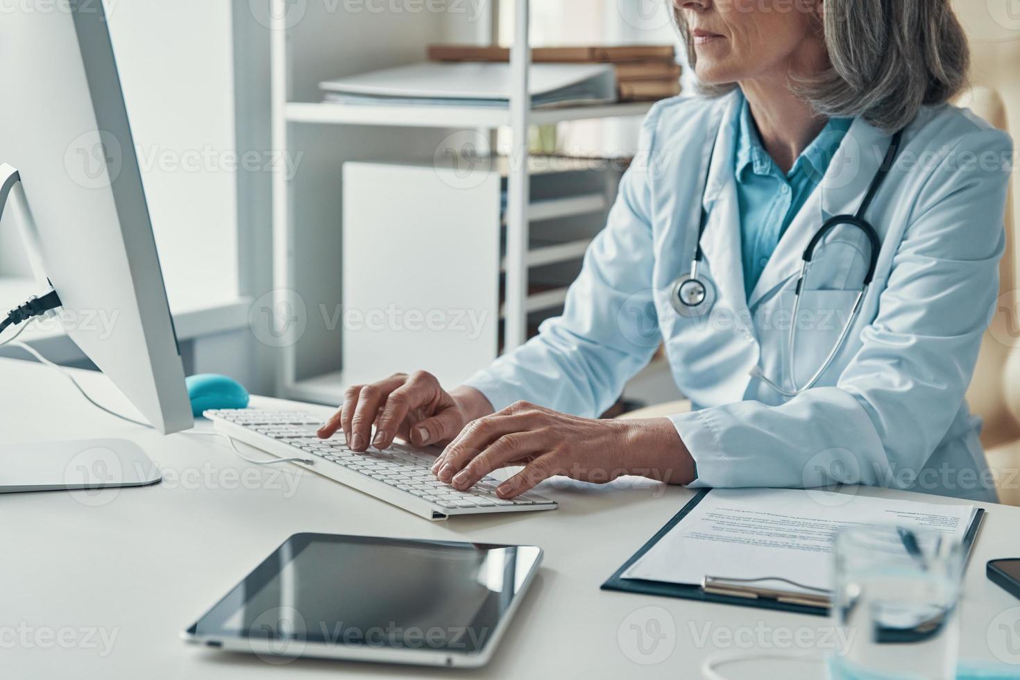 Close up of female doctor in white lab coat working using computer while sitting in her office photo