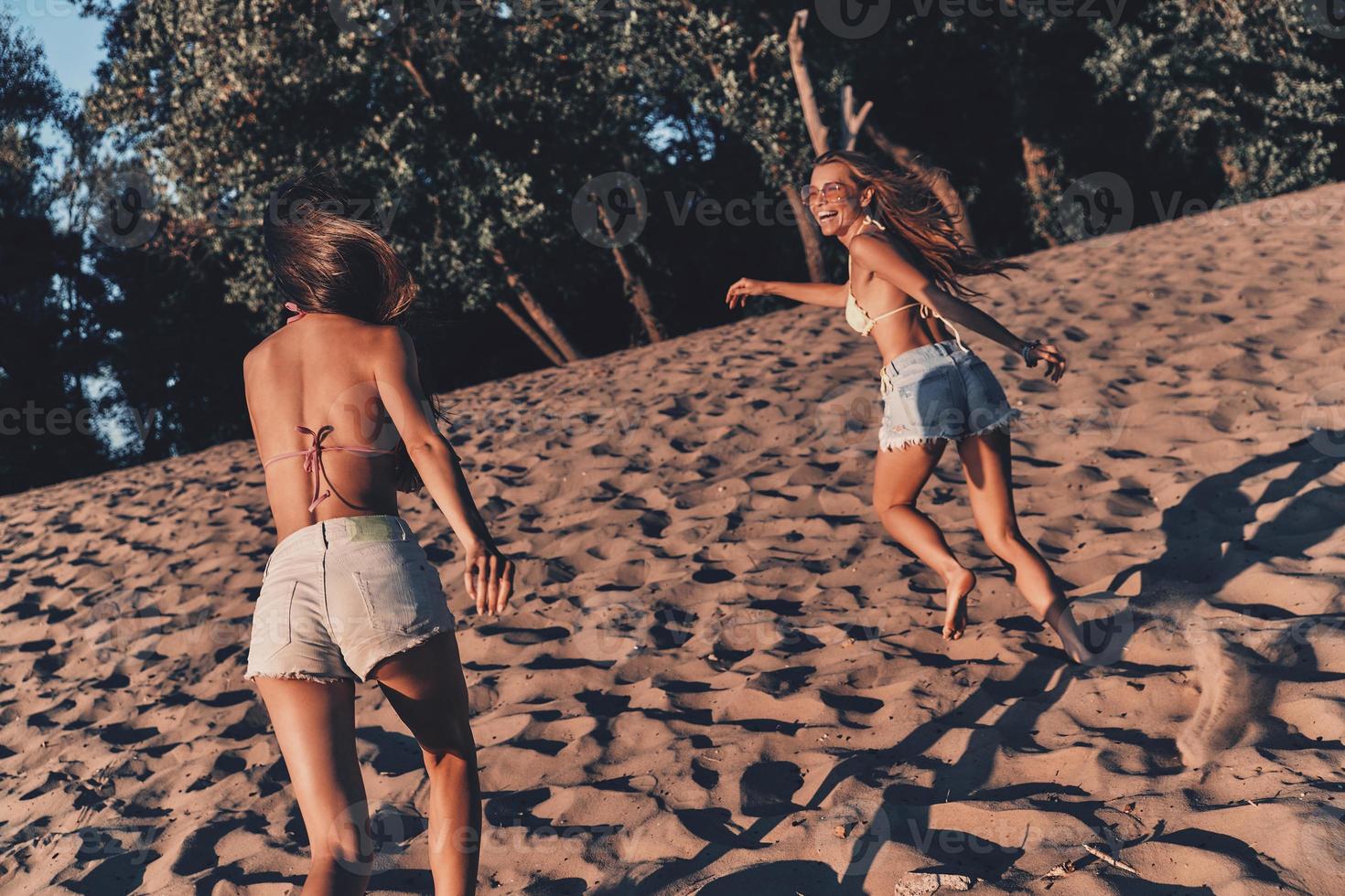 dos atractivas mujeres jóvenes en pantalones cortos y trajes de baño sonriendo mientras corren en la playa foto