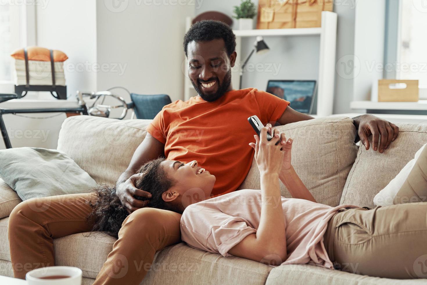 Young African woman using smart phone and smiling while spending time at home with her boyfriend photo