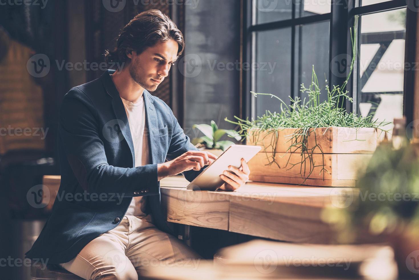 Working just from cafe. Confident young man in smart casual wear working on digital tablet while sitting near window in cafe photo