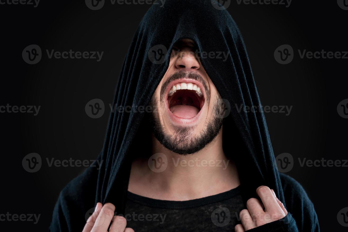 Unleashed emotions. Young man covering his face by black hood and shouting while standing against black background photo