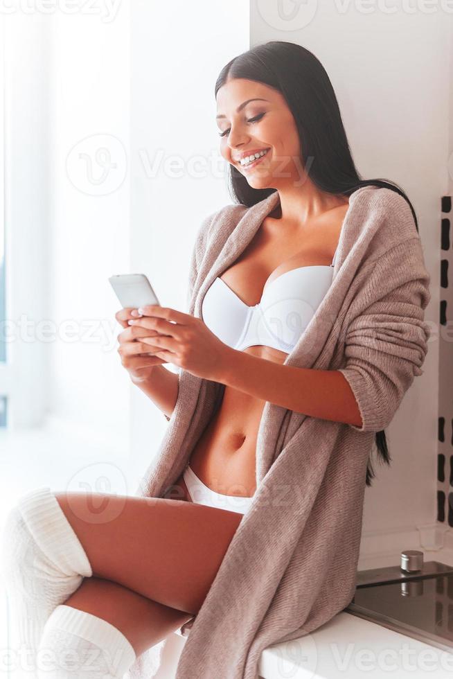 Lovely message from him. Beautiful young woman in lingerie and sweater holding mobile phone and looking at it with smile while sitting on the kitchen counter at home photo