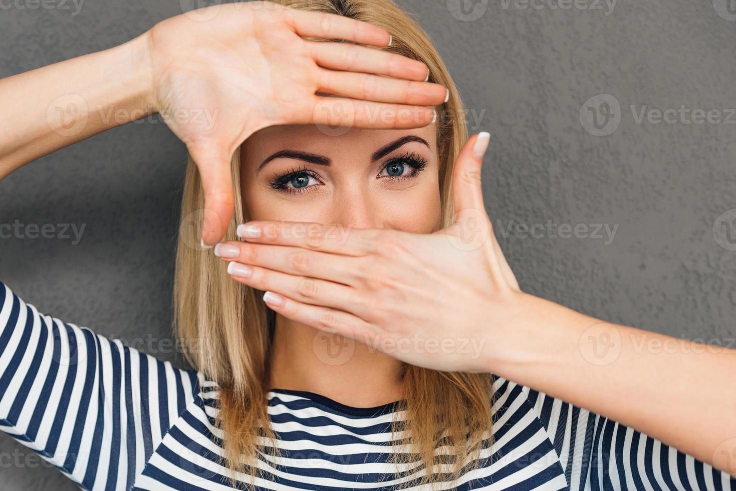 Focus on my eyes Beautiful young woman making finger frame and looking at camera while standing against grey background photo