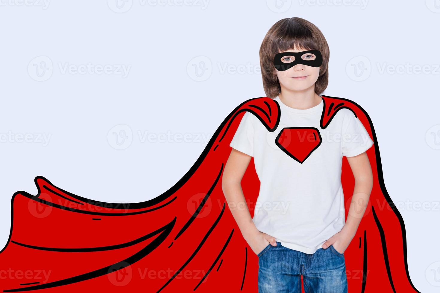 Like a hero. Cheerful boy holding hands in pockets and looking like superhero in his drawn cape while standing against white background photo