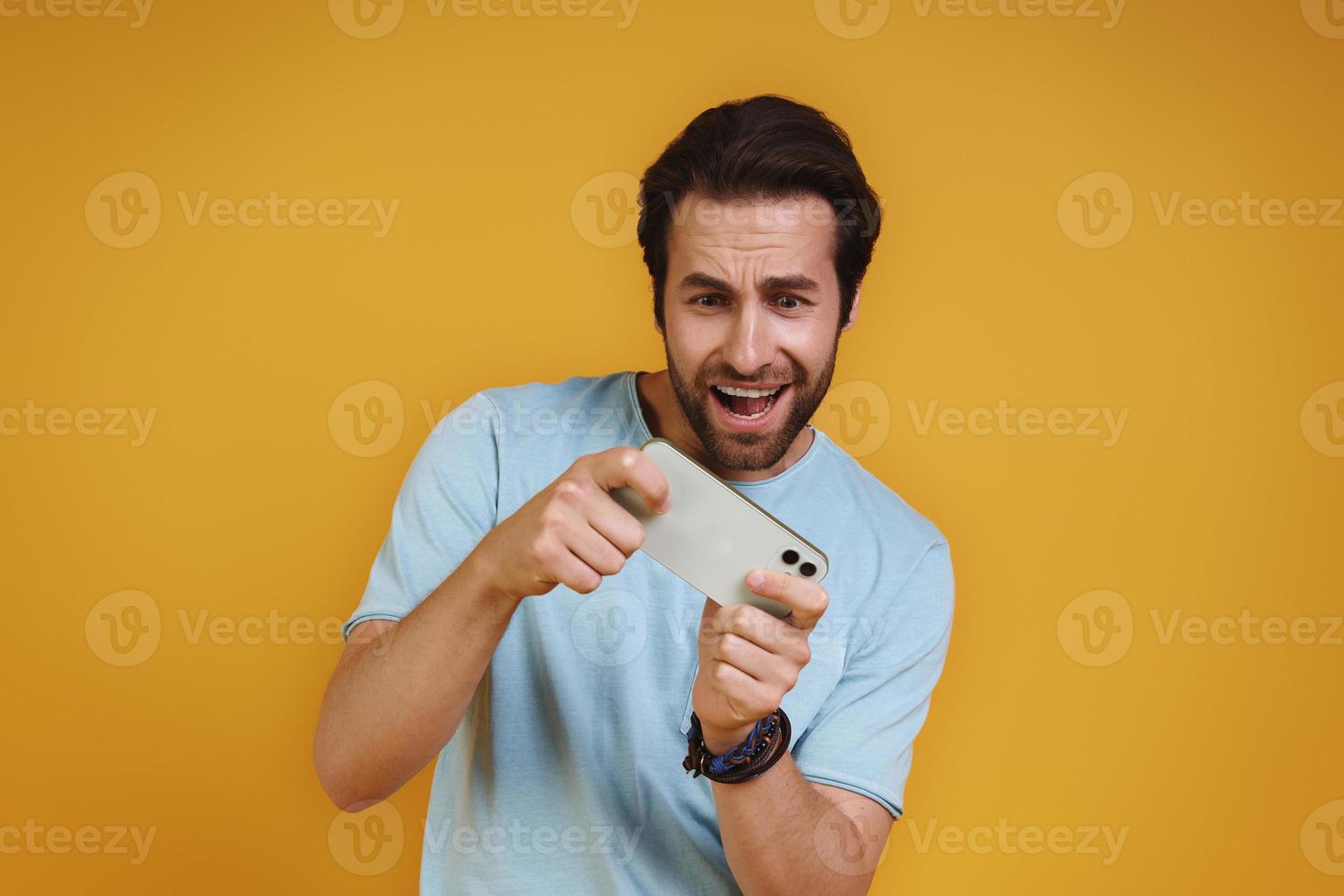 Happy young man in casual clothing playing games on smart phone and smiling photo