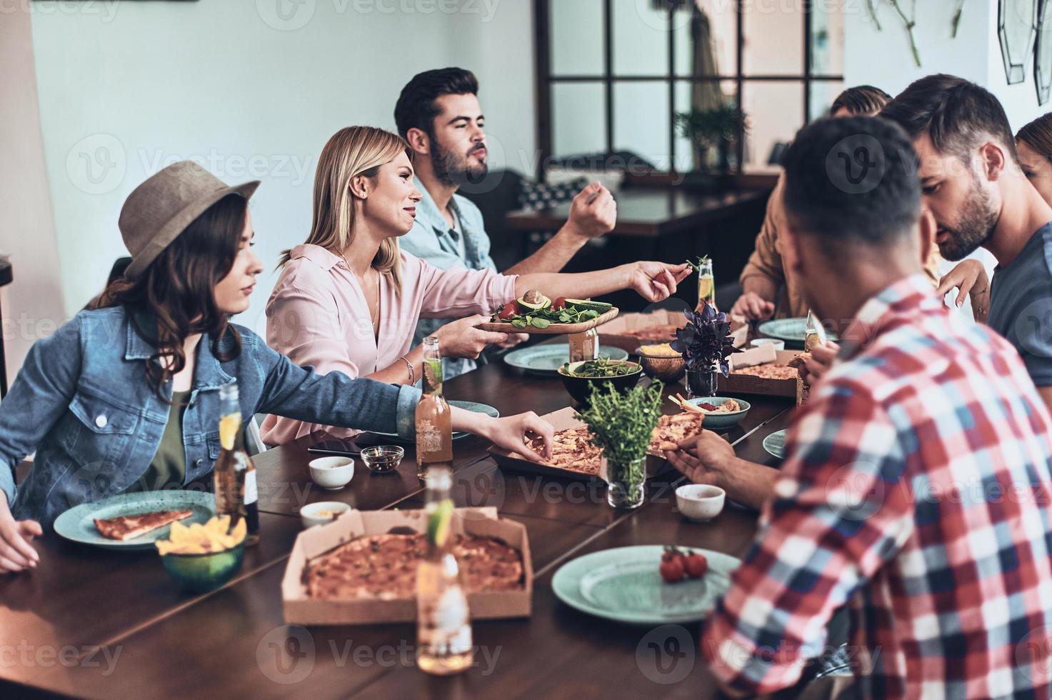 La vida es mejor con amigos. grupo de jóvenes con ropa informal comiendo y sonriendo mientras cenan en el interior foto