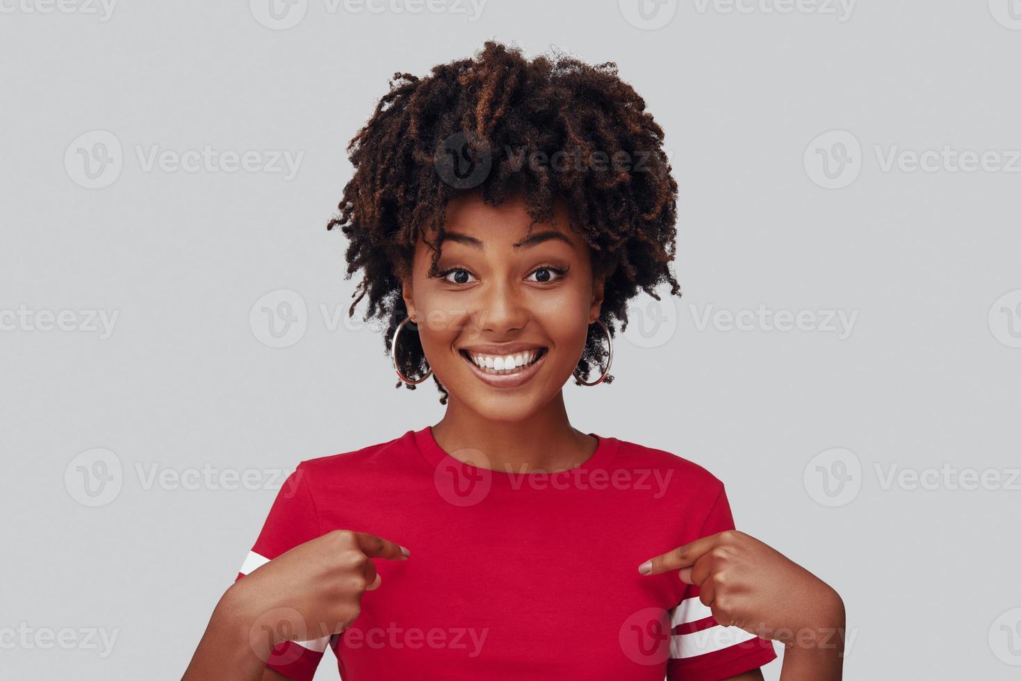 Attractive young African woman looking at camera with smile and pointing at herself while standing against grey background photo