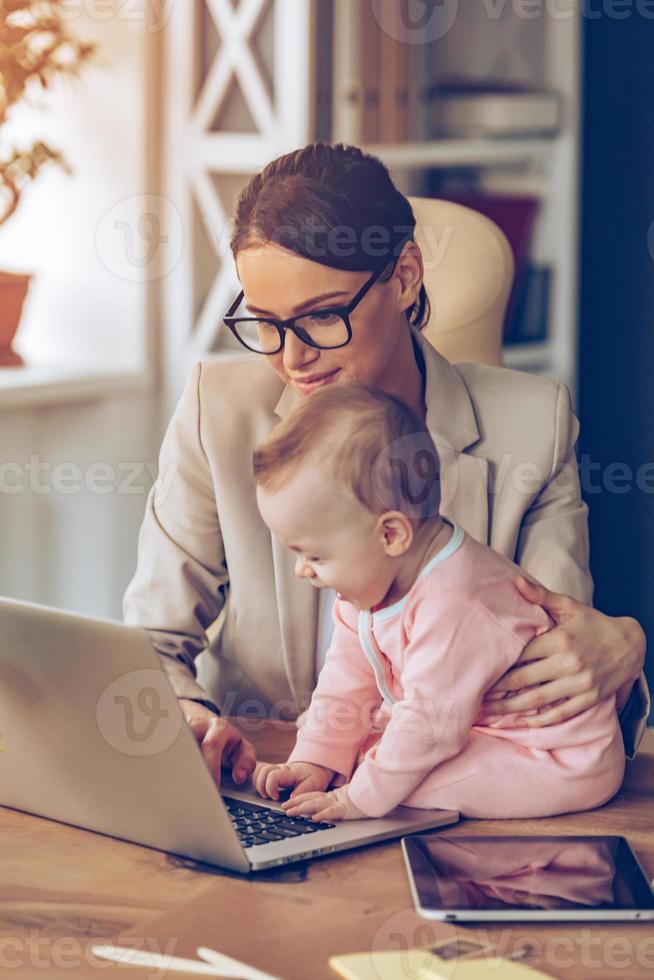 Helping mom. Little baby girl using laptop while sitting on office desk with her mother in office photo