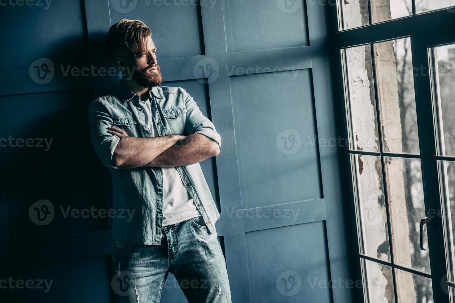 pensando en... un apuesto joven barbudo con pantalones vaqueros claros que mantiene los brazos cruzados y mira hacia otro lado mientras está de pie cerca de la ventana foto