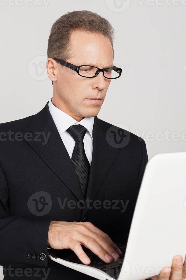 Businessman with laptop. Confident mature man in formalwear working on laptop while standing against grey background photo