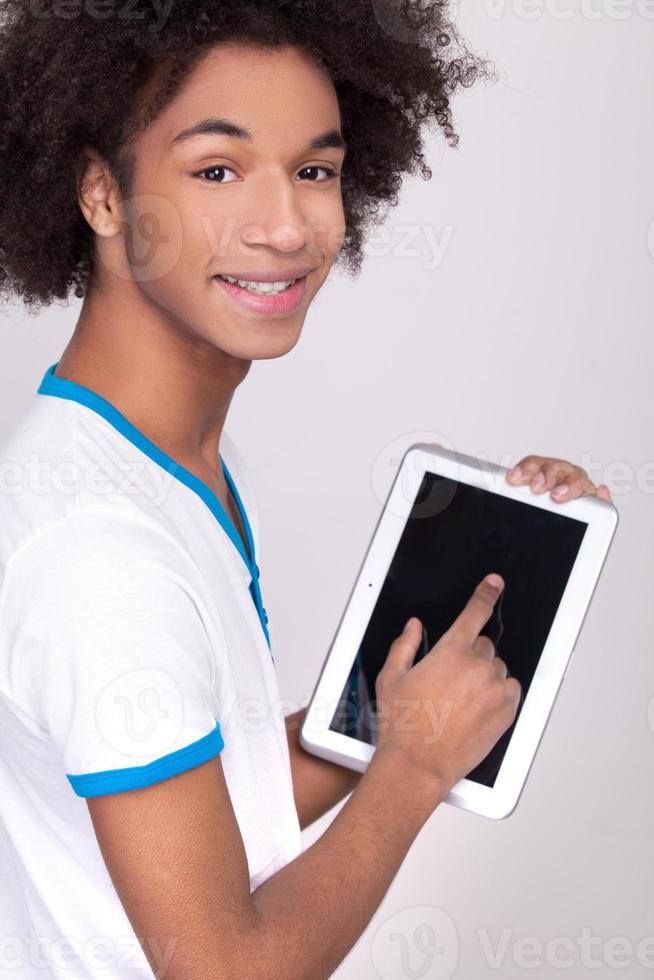 Working on digital tablet. Rear view of cheerful African teenager working on digital tablet and looking over shoulder while standing isolated on grey background photo