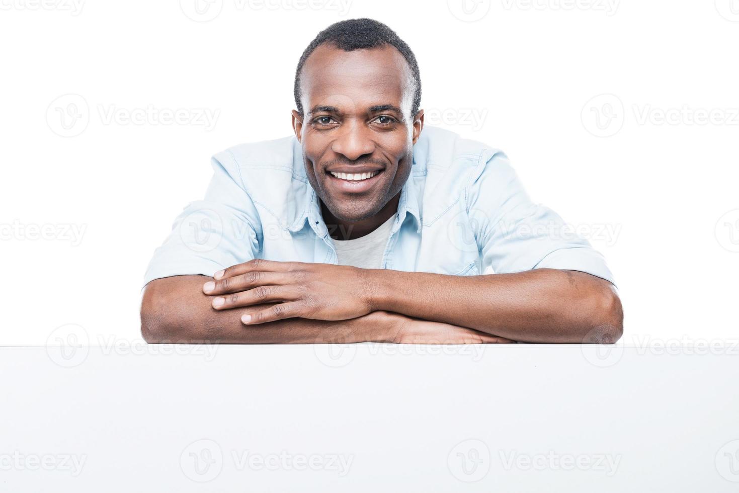 encantador guapo. hombre africano guapo con camisa azul apoyado en el espacio de la copia y sonriendo mientras está de pie contra el fondo blanco foto