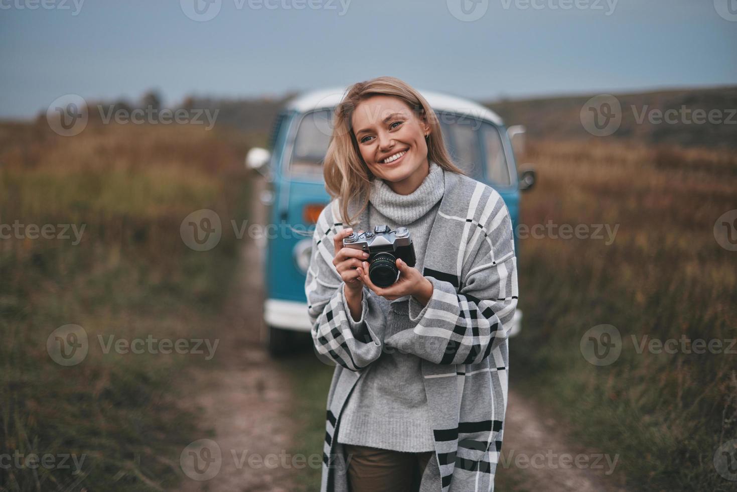 joven fotógrafo. atractiva mujer joven sosteniendo una cámara digital y sonriendo mientras está de pie al aire libre con la mini furgoneta azul de estilo retro en el fondo foto